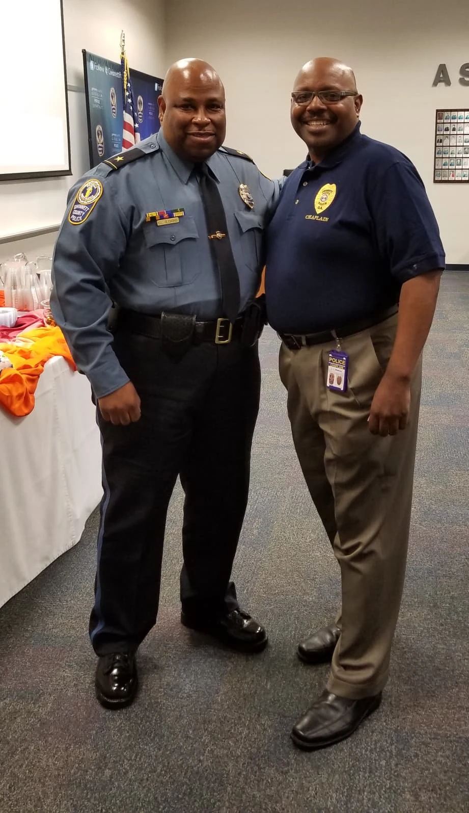 Two men standing together at a community event, one in a police uniform and one in a chaplain polo shirt.