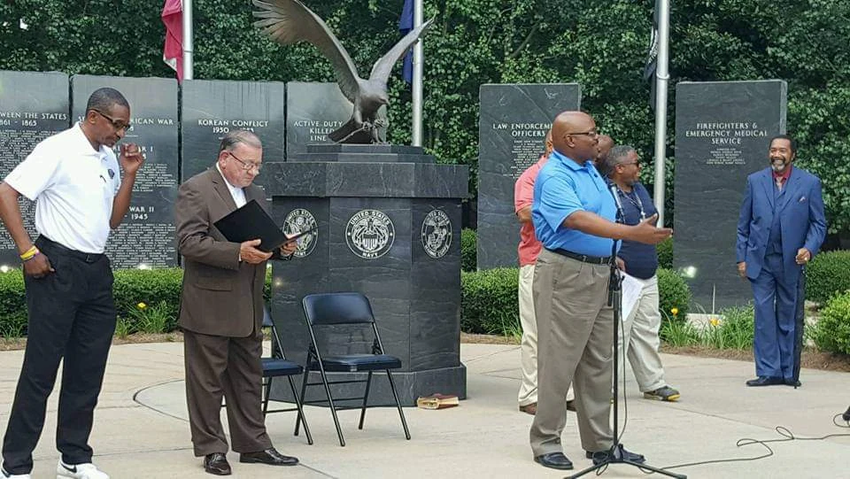 A speaker at a microphone with community members gathered beside a memorial.