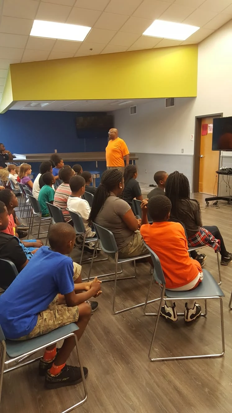 A speaker addressing a group of young people seated in a community room.